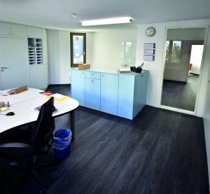 Gemeindebüro Die Aufnahme zeigt den Innenraum eines hellen und modern eingerichteten Gemeindebüros der Evangelischen Kirchengemeinde Hochdahl, das einen funktionalen und aufgeräumten Eindruck macht. Das Licht kommt von oben durch eine Deckenleuchte und ein Fenster, was den Raum gut ausleuchtet. Boden und Wände Der Boden ist mit einem dunklen Holzimitat-Laminat ausgelegt, der eine deutliche Maserung zeigt und dem Raum einen modernen Kontrast zu den hellen Wänden verleiht. Die Wände sind durchweg in einem sauberen Weiß gehalten. Vordergrund: Arbeitsbereich Im linken Vordergrund ist ein großer, heller Bürotisch zu sehen, der eine geschwungene oder L-förmige Platte hat. Auf dem Tisch liegen verschiedene Büromaterialien wie Blätter, Stifte und möglicherweise ein Telefon, was auf einen aktiven Arbeitsplatz hindeutet. Davor steht ein schwarzer Bürostuhl mit Armlehnen auf Rollen, der zum Tisch hin ausgerichtet ist. Unter dem Tisch, nahe dem Stuhl, steht ein kleiner, blauer Papierkorb mit einer transparenten blauen Mülltüte. Mitte: Aktenschränke und Thekenbereich Die Mitte des Raumes wird von einem auffälligen, türkisfarbenen oder hellblauen Schrankelement dominiert, das als Theke oder Ablagefläche dient. Dieses Möbelstück hat mehrere Schranktüren und Schubladen auf der Vorderseite, was auf Stauraum hindeutet. Auf der Oberfläche dieses Schrankelements liegen weitere Büromaterialien, möglicherweise ein Ordner und ein Stiftehalter. Dahinter, an der Wand, ist ein großes, vertikales Fenster zu sehen, durch das Tageslicht einfällt und einen Blick nach draußen auf andere Gebäude und Grünflächen erlaubt. Hintergrund: Schränke, Türen und Nebenräume An der linken Wand, im Hintergrund, erstreckt sich eine lange Reihe von weißen Hängeschränken bis zur Decke, die zusätzlichen Stauraum bieten. Einige dieser Schränke haben offene Fächer mit sortierten Ablagen, was auf eine organisierte Ablage von Dokumenten hindeutet. Rechts vom türkisfarbenen Schrankelement ist eine helle, schlichte Bürotür zu erkennen, die leicht geöffnet ist. Durch den Türrahmen hindurch ist ein weiterer Raum sichtbar, der ebenfalls den dunklen Boden und helle Wände aufweist. In diesem Nebenraum ist ein auffälliger roter oder orangefarbener Stuhl oder Sessel zu sehen, der einen Farbakzent setzt. An der Wand über dem Schrankelement hängt eine runde Wanduhr mit weißen Ziffernblättern und schwarzen Zeigern. Daneben hängt ein Wandkalender mit drei Monatsübersichten untereinander. Decke und Beleuchtung Die Decke ist weiß und schlicht. Eine lange, rechteckige Deckenleuchte mit Leuchtstoffröhren sorgt für zusätzliche helle Beleuchtung im Raum. Insgesamt vermittelt das Bild den Eindruck eines gut organisierten, hellen und funktionalen Büroumfelds.