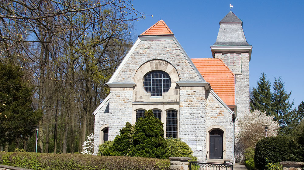 Neanderkirche in Erkrath-Hochdahl Das Bild zeigt die Neanderkirche in Erkrath-Hochdahl. Das Gebäude besteht aus hellem Naturstein und hat ein steiles, orange-rotes Ziegeldach. Links und rechts neben dem Haupteingang ragen Mauern hoch, in deren Mitte ein großes, rundbogiges Kirchenfenster eingelassen ist. Darunter befinden sich zwei kleinere, rechteckige Fenster. Der Haupteingang ist ein dunkles Holztor auf der rechten Seite. Rechts vom Hauptbau erhebt sich ein quadratischer Kirchturm mit grauem Schieferdach, das in einer spitzen Form ausläuft. Auf der Spitze sitzt ein Wetterhahn. Die Kirche ist von Bäumen und Büschen eingerahmt. Im Vordergrund sieht man grüne Hecken und eine kleine Mauer, die das Grundstück abgrenzt. Der Himmel ist klar und blau, sodass das helle Gestein der Kirche besonders auffällt.
