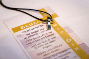 Konfirmation Das Foto zeigt ein silbernes Kreuz an einer schwarzen Kordel, das auf einem Blatt Papier liegt. Das Blatt ist gestaltet wie eine kleine Karte mit einem Segenstext. Am linken oberen Rand steht das Wort „SEGEN“ in weißen Großbuchstaben auf einem gelben Hintergrund. Rechts davon ist senkrecht das Wort „MIT DIR“ ebenfalls auf gelbem Hintergrund gedruckt. Der Text auf der Karte lautet auszugsweise: „Segen sei mit Dir, der Segen des strahlenden Lichtes. Licht sei um Dich her und in Deinem Herzen. Gott sei Schutz und Schirm vor allem Argen …“ Die Karte liegt auf einem weißen Untergrund, vermutlich einer Tischdecke. Das Kreuz wirkt glänzend und modern, eher schlicht gestaltet. Das Gesamtbild vermittelt eine ruhige, feierliche Stimmung, passend zu einem religiösen Anlass wie Konfirmation.