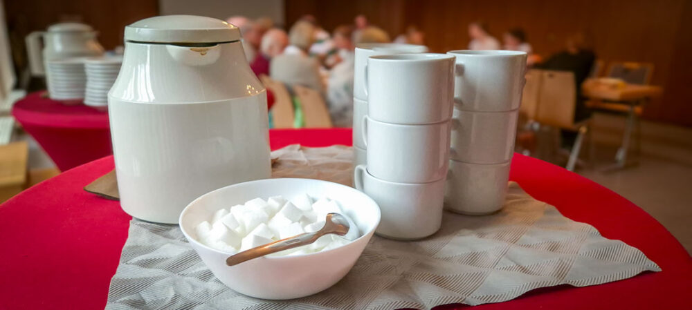 Das Bild zeigt einen Ausschnitt eines Kaffeetrinkens bei der Evangelischen Kirchengemeinde Hochdahl, wobei der Fokus auf dem Service-Bereich in der vorderen Mitte liegt. Im Vordergrund auf einem roten, runden Tisch steht eine kleine Arrangement für Kaffee. Zentral positioniert ist eine große, weiße Kaffeekanne (oder Thermoskanne) mit Deckel. Rechts davon sind zwei Stapel weißer Tassen zu sehen, jeweils drei Tassen hoch gestapelt. Davor, mittig, steht eine kleine weiße Schale, gefüllt mit weißen Zuckerwürfeln. Ein kleiner Löffel liegt in der Schale. Das gesamte Arrangement steht auf einer quadratischen Serviette oder einem kleinen Tuch mit einem subtilen, geometrischen Muster in Grau- und Beigetönen. Links von der Kanne ist ein weiterer Stapel kleiner, weißer Teller und eine kleinere Kanne oder Teekanne erkennbar. Der Hintergrund ist unscharf gehalten, was die Aufmerksamkeit auf das Arrangement lenkt, aber es sind mehrere Personen zu erkennen, die an Tischen sitzen und sich unterhalten, was auf eine Veranstaltung oder ein geselliges Beisammensein hindeutet. Die Beleuchtung ist hell, was auf einen gut beleuchteten Innenraum schließen lässt.