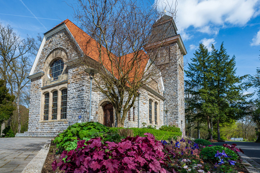 Evangelische Neanderkirche in Hochdahl aus grauem Naturstein mit rotem Ziegeldach, davor ein Beet mit roten und violetten Pflanzen und ein kahler Baum, bei blauem Himmel.